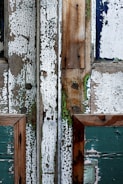 A close-up view of weathered wooden surfaces, featuring chipped and peeling paint. The wood is composed of several panels with various shades of white, green, and natural wood tones, showing significant texture and evidence of decay. The paint appears aged, cracked, and worn, revealing underlying layers and hints of mossy green.