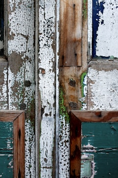 A close-up view of weathered wooden surfaces, featuring chipped and peeling paint. The wood is composed of several panels with various shades of white, green, and natural wood tones, showing significant texture and evidence of decay. The paint appears aged, cracked, and worn, revealing underlying layers and hints of mossy green.