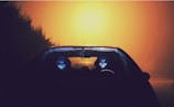 Two figures wearing alien masks are seated inside a car, viewed from the front. The setting is illuminated by an eerie orange-yellow light creating a foggy and mysterious atmosphere.
