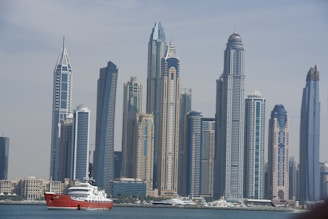 A skyline featuring modern skyscrapers with distinctive architectural designs, set against a clear sky. In front of the skyline, a red and white yacht is visible on the water.