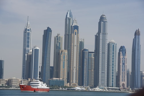 A skyline featuring modern skyscrapers with distinctive architectural designs, set against a clear sky. In front of the skyline, a red and white yacht is visible on the water.
