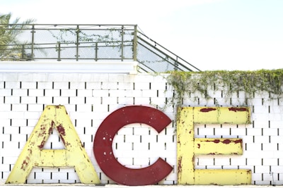 Large, vintage-style letters spell out 'ACE' against a white brick wall. The letters are weathered, with visible rust on the red 'C' and yellow 'A' and 'E'. Green vines drape over the top of the wall, adding a natural element to the scene. A metal fence and palm tree can be seen in the background.