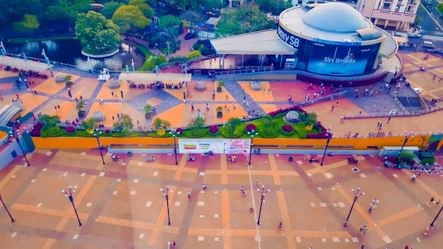 An aerial view of a bustling urban area featuring a large plaza with people walking around. There's a round building with signage, surrounded by greenery and a small body of water with pathways. The plaza is adorned with geometric patterns and lampposts.