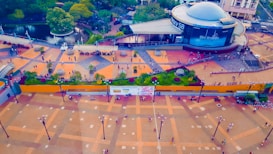 An aerial view of a bustling urban area featuring a large plaza with people walking around. There's a round building with signage, surrounded by greenery and a small body of water with pathways. The plaza is adorned with geometric patterns and lampposts.
