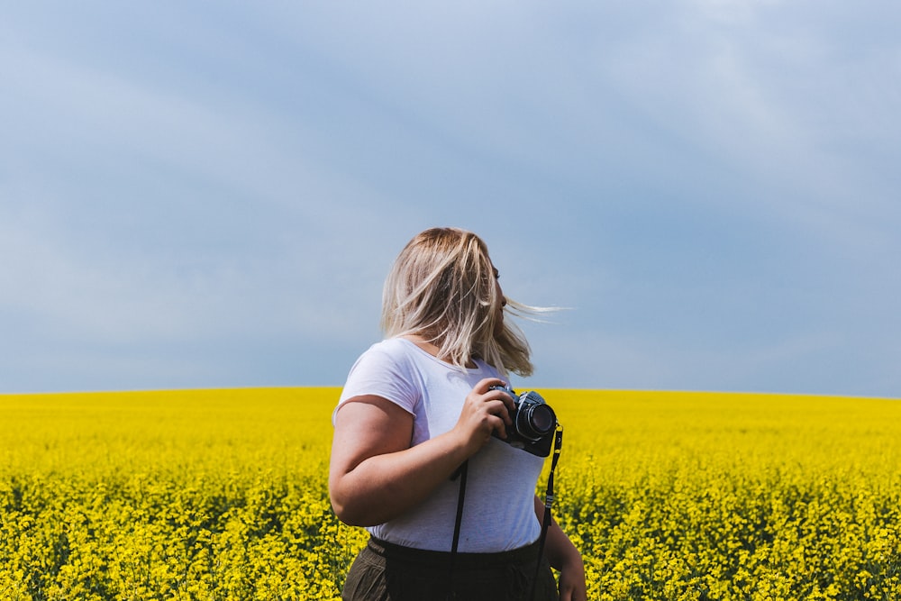 mulher segurando a câmera DSLR em pé na frente do campo de flores amarelas