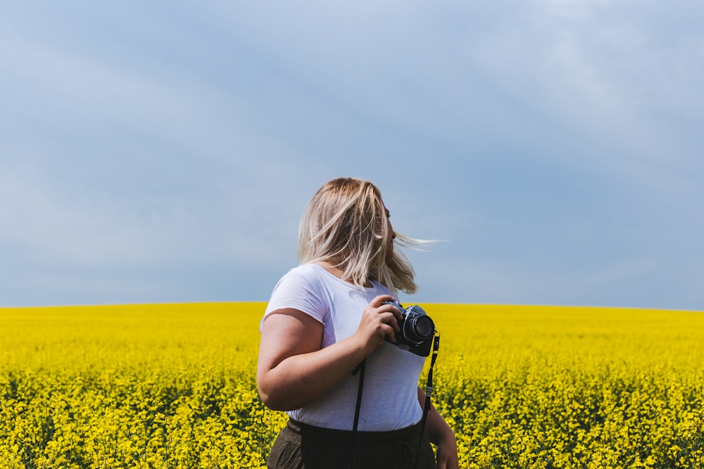 mujer sosteniendo una cámara DSLR de pie frente a un campo de flores amarillas