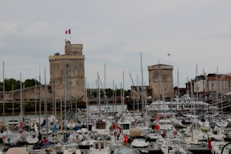 A harbor filled with numerous sailboats and yachts, with two large stone towers in the background topped by French flags. The towers appear historic, suggesting a blend of medieval and maritime elements. Surrounding the harbor are buildings with red and white facades.