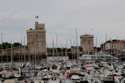 A harbor filled with numerous sailboats and yachts, with two large stone towers in the background topped by French flags. The towers appear historic, suggesting a blend of medieval and maritime elements. Surrounding the harbor are buildings with red and white facades.