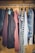 A collection of various clothing items hanging neatly on a metal rod inside a wooden wardrobe. The clothing includes jackets, shirts, and tops with different colors and patterns such as stripes and plaid.