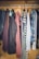 A collection of various clothing items hanging neatly on a metal rod inside a wooden wardrobe. The clothing includes jackets, shirts, and tops with different colors and patterns such as stripes and plaid.