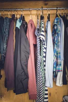 A collection of various clothing items hanging neatly on a metal rod inside a wooden wardrobe. The clothing includes jackets, shirts, and tops with different colors and patterns such as stripes and plaid.