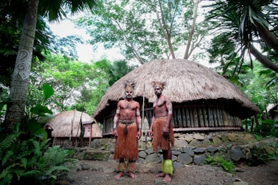 Two people are standing in front of a traditional thatched-roof hut surrounded by lush greenery. They are wearing cultural attire and have body paint and accessories that reflect traditional designs. The background features dense trees and plants, creating a natural setting.