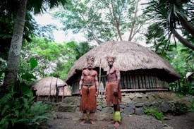 Two people are standing in front of a traditional thatched-roof hut surrounded by lush greenery. They are wearing cultural attire and have body paint and accessories that reflect traditional designs. The background features dense trees and plants, creating a natural setting.