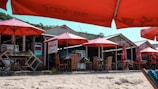 A beachside scene with several wooden huts and shaded seating areas. The seating is covered by large red umbrellas, and the sandy beach area is in the foreground. The huts have signboards, and there is a small path or walkway in front of them. There are a few people visible around the huts.