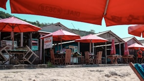 A beachside scene with several wooden huts and shaded seating areas. The seating is covered by large red umbrellas, and the sandy beach area is in the foreground. The huts have signboards, and there is a small path or walkway in front of them. There are a few people visible around the huts.