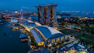 Aerial view of a cityscape at dusk, featuring a large, illuminated resort complex with three interconnected towers and a boat-shaped structure on top. Nearby, a prominent ferris wheel glows with vibrant colors. The area is surrounded by water, with numerous ships dotting the horizon. Lush greenery and modern architectural structures are visible in the vicinity.