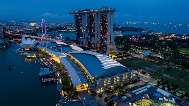 Aerial view of a cityscape at dusk, featuring a large, illuminated resort complex with three interconnected towers and a boat-shaped structure on top. Nearby, a prominent ferris wheel glows with vibrant colors. The area is surrounded by water, with numerous ships dotting the horizon. Lush greenery and modern architectural structures are visible in the vicinity.