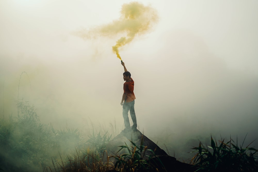 Homme debout sur une formation rocheuse tenant une fusée éclairante jaune entourée de brouillards