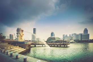 A cityscape with a prominent Merlion statue on the left corner, spouting water into Marina Bay. The background features modern skyscrapers and a domed building near a waterfront promenade. The sky is partly cloudy, casting a soft light over the scene.
