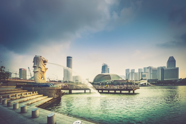 A cityscape with a prominent Merlion statue on the left corner, spouting water into Marina Bay. The background features modern skyscrapers and a domed building near a waterfront promenade. The sky is partly cloudy, casting a soft light over the scene.