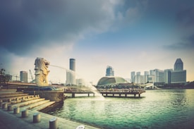 A cityscape with a prominent Merlion statue on the left corner, spouting water into Marina Bay. The background features modern skyscrapers and a domed building near a waterfront promenade. The sky is partly cloudy, casting a soft light over the scene.