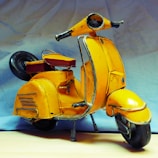 A vintage toy scooter with a bright yellow body and black wheels is positioned against a soft, light blue background. The scooter features a red seat and silver detailing, giving it a classic and nostalgic appearance.