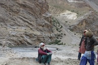 A rugged, mountainous landscape with towering rock formations. Three individuals, dressed in traditional attire and head coverings, are positioned on a slope. Two are standing, while one is seated on the dirt path. A few black animals, possibly yaks or cows, can be seen in the distance near a shallow body of water.