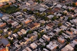 An aerial view of an urban neighborhood featuring tightly packed residential buildings, roadways, and interspersed green areas. The scene captures a variety of architecture styles with flat roofs, and multiple-story structures. Trees line the streets, creating patches of green amid the predominantly beige and gray buildings.