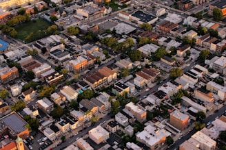 An aerial view of an urban neighborhood featuring tightly packed residential buildings, roadways, and interspersed green areas. The scene captures a variety of architecture styles with flat roofs, and multiple-story structures. Trees line the streets, creating patches of green amid the predominantly beige and gray buildings.