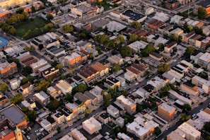 An aerial view of an urban neighborhood featuring tightly packed residential buildings, roadways, and interspersed green areas. The scene captures a variety of architecture styles with flat roofs, and multiple-story structures. Trees line the streets, creating patches of green amid the predominantly beige and gray buildings.