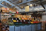 A neatly organized section of a grocery store featuring an array of colorful fruits and vegetables displayed in open bins. Above the produce, there are woven baskets. In the background, shelves are stocked with various packaged goods and boxes. There are some flower arrangements on the counter to the left, and the lighting is bright with multiple ceiling lights illuminating the area. The store has a modern industrial design with exposed ducts and metal framework.