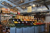 A neatly organized section of a grocery store featuring an array of colorful fruits and vegetables displayed in open bins. Above the produce, there are woven baskets. In the background, shelves are stocked with various packaged goods and boxes. There are some flower arrangements on the counter to the left, and the lighting is bright with multiple ceiling lights illuminating the area. The store has a modern industrial design with exposed ducts and metal framework.