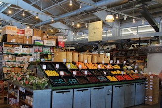 A neatly organized section of a grocery store featuring an array of colorful fruits and vegetables displayed in open bins. Above the produce, there are woven baskets. In the background, shelves are stocked with various packaged goods and boxes. There are some flower arrangements on the counter to the left, and the lighting is bright with multiple ceiling lights illuminating the area. The store has a modern industrial design with exposed ducts and metal framework.