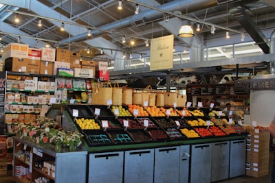 A neatly organized section of a grocery store featuring an array of colorful fruits and vegetables displayed in open bins. Above the produce, there are woven baskets. In the background, shelves are stocked with various packaged goods and boxes. There are some flower arrangements on the counter to the left, and the lighting is bright with multiple ceiling lights illuminating the area. The store has a modern industrial design with exposed ducts and metal framework.