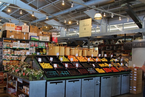 A neatly organized section of a grocery store featuring an array of colorful fruits and vegetables displayed in open bins. Above the produce, there are woven baskets. In the background, shelves are stocked with various packaged goods and boxes. There are some flower arrangements on the counter to the left, and the lighting is bright with multiple ceiling lights illuminating the area. The store has a modern industrial design with exposed ducts and metal framework.