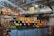 A neatly organized section of a grocery store featuring an array of colorful fruits and vegetables displayed in open bins. Above the produce, there are woven baskets. In the background, shelves are stocked with various packaged goods and boxes. There are some flower arrangements on the counter to the left, and the lighting is bright with multiple ceiling lights illuminating the area. The store has a modern industrial design with exposed ducts and metal framework.
