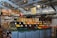 A neatly organized section of a grocery store featuring an array of colorful fruits and vegetables displayed in open bins. Above the produce, there are woven baskets. In the background, shelves are stocked with various packaged goods and boxes. There are some flower arrangements on the counter to the left, and the lighting is bright with multiple ceiling lights illuminating the area. The store has a modern industrial design with exposed ducts and metal framework.