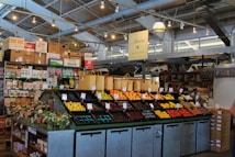 A neatly organized section of a grocery store featuring an array of colorful fruits and vegetables displayed in open bins. Above the produce, there are woven baskets. In the background, shelves are stocked with various packaged goods and boxes. There are some flower arrangements on the counter to the left, and the lighting is bright with multiple ceiling lights illuminating the area. The store has a modern industrial design with exposed ducts and metal framework.