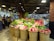 A supermarket interior featuring several baskets filled with fruits, predominantly apples. The apples are wrapped with pink protective foam. Numerous people are shopping, browsing the displays under artificial lighting that highlights the produce section. In the background, preserved fruit sections and other food items are visible on shelves.