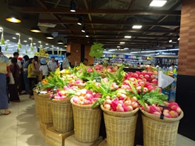 A supermarket interior featuring several baskets filled with fruits, predominantly apples. The apples are wrapped with pink protective foam. Numerous people are shopping, browsing the displays under artificial lighting that highlights the produce section. In the background, preserved fruit sections and other food items are visible on shelves.