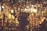 A dimly lit market stall is filled with an array of copper pots, pans, and other metallic items hanging from above and stacked on shelves. Warm light bulbs cast a soft glow over the metallic surfaces, enhancing the rich, golden and copper hues. In the center, a person stands amidst the hanging items, creating a cozy and busy atmosphere.