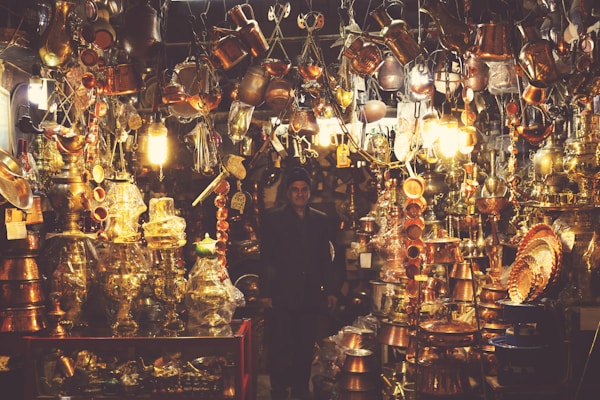 A dimly lit market stall is filled with an array of copper pots, pans, and other metallic items hanging from above and stacked on shelves. Warm light bulbs cast a soft glow over the metallic surfaces, enhancing the rich, golden and copper hues. In the center, a person stands amidst the hanging items, creating a cozy and busy atmosphere.