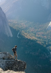 man standing on grey rock on cliff with overview of forest during daytime