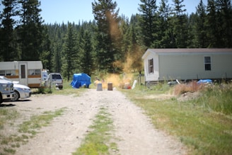 A gravel driveway leads through a rural area with scattered objects and vehicles. On the left, an RV and a few cars are parked near some trees. A blue tarp covers an item in the background, and several trash barrels are placed along the pathway. A cloud of orange dust or smoke rises in the center of the image. On the right, a small mobile home is visible, surrounded by grass and trees.
