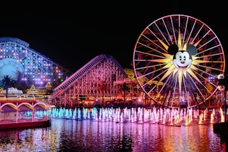 A vibrant amusement park scene at night with a large Ferris wheel featuring a popular cartoon character's face. The area is illuminated with multicolored lights, reflecting on the water. A roller coaster is visible in the background with colorful lights outlining its structure. Fountains in the foreground are lit up in various colors, creating a lively and festive atmosphere.