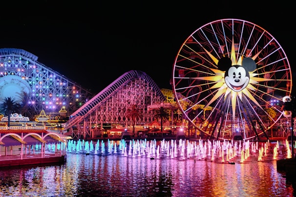 A vibrant amusement park scene at night with a large Ferris wheel featuring a popular cartoon character's face. The area is illuminated with multicolored lights, reflecting on the water. A roller coaster is visible in the background with colorful lights outlining its structure. Fountains in the foreground are lit up in various colors, creating a lively and festive atmosphere.