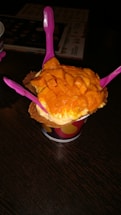 A cup of ice cream topped with fresh mango chunks is presented on a dark wooden table. Three bright pink plastic spoons are inserted into the ice cream, and the treat is placed in a waffle cone bowl within a colorful paper cup.