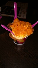 A cup of ice cream topped with fresh mango chunks is presented on a dark wooden table. Three bright pink plastic spoons are inserted into the ice cream, and the treat is placed in a waffle cone bowl within a colorful paper cup.