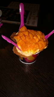 A cup of ice cream topped with fresh mango chunks is presented on a dark wooden table. Three bright pink plastic spoons are inserted into the ice cream, and the treat is placed in a waffle cone bowl within a colorful paper cup.