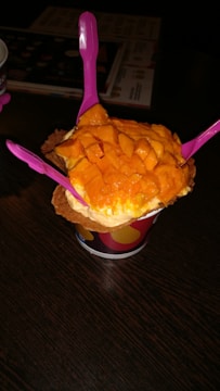 A cup of ice cream topped with fresh mango chunks is presented on a dark wooden table. Three bright pink plastic spoons are inserted into the ice cream, and the treat is placed in a waffle cone bowl within a colorful paper cup.